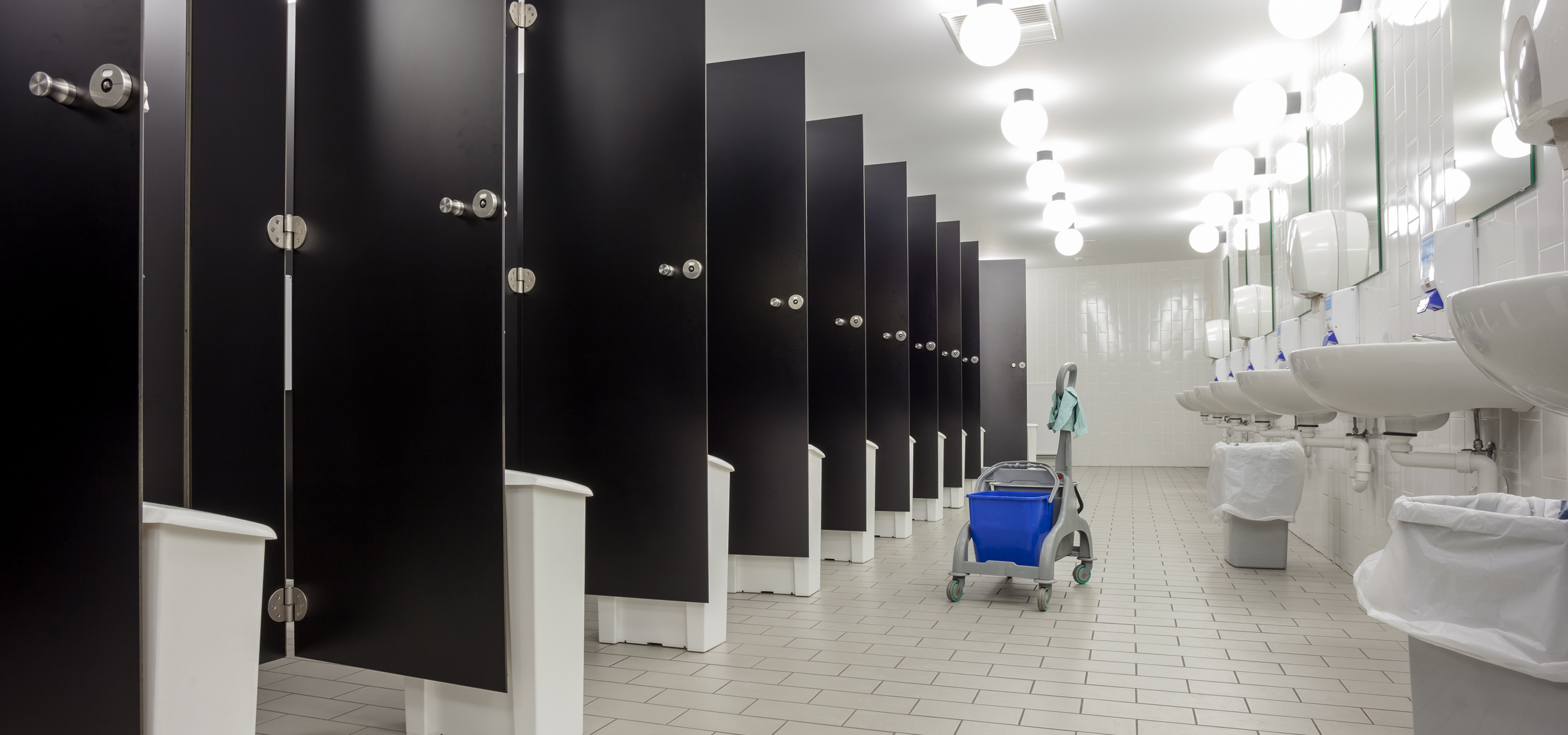 A perspective photo of a very long and very white bathroom with sinks and mirrors on one side, a severe number of globe lights overhead at equal intervals, and white tile on the floor in brick pattern with dark grout. Opposite the wall of sinks and mirrors are a long row of black toilet partitions. Every partition door is propped open by a white waste basket, creating a repeating effect. A single blue and grey mop bucket sits in between the two sides and offers the only spot of color in the room.